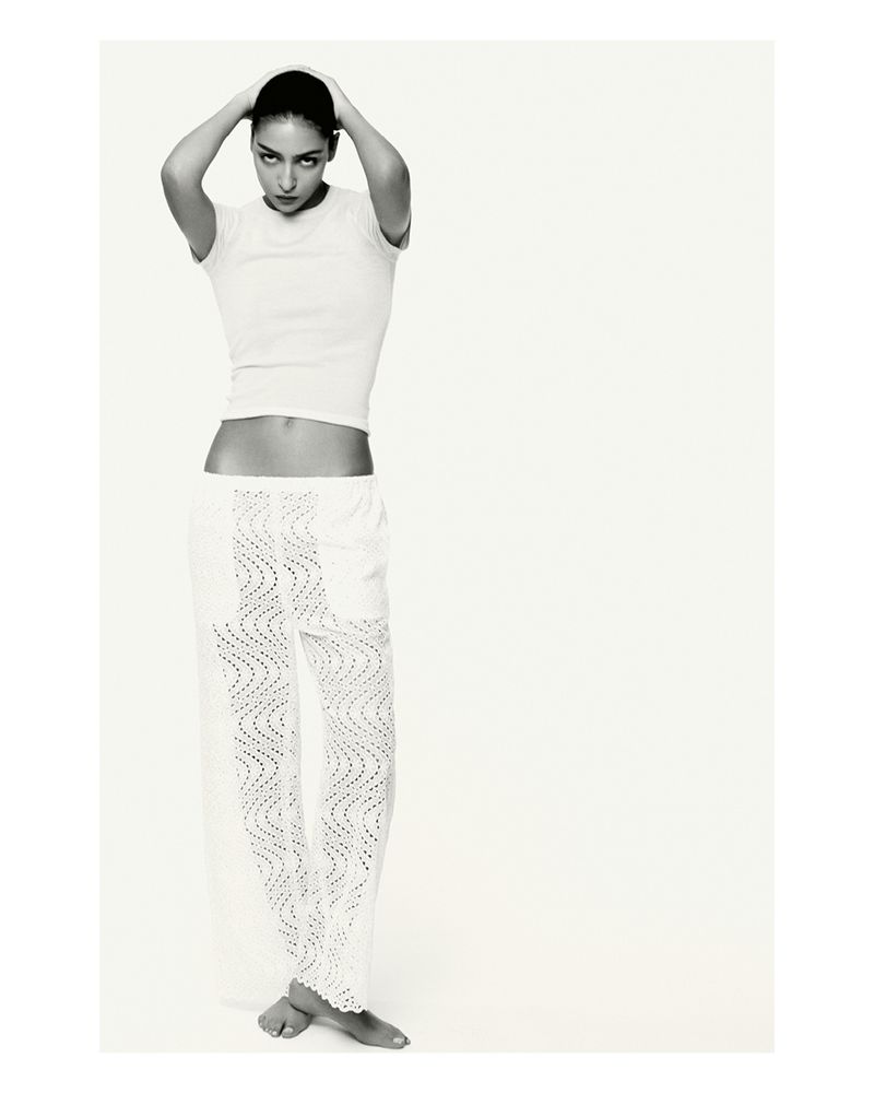 Black and white portrait of an adult woman with hands behind her head, wearing a white t-shirt and lace pants, against a neutral backdrop.