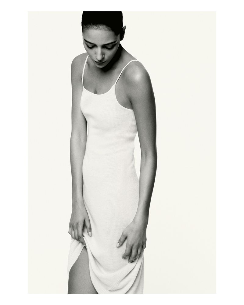 Elegant portrait of a slender adult woman in a white knit dress, looking down against a neutral background in a studio environment.