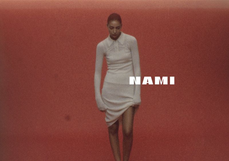 A woman in a white dress stands in front of a terracotta background, with the text “Nami” overlaid across the center of the frame.