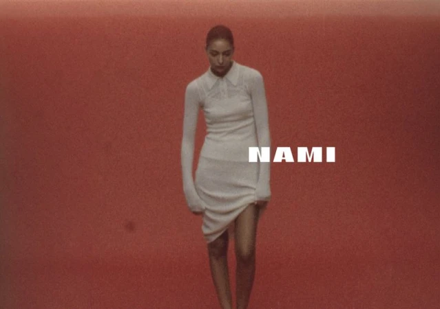 A woman in a white dress stands in front of a terracotta background, with the text “Nami” overlaid across the center of the frame.