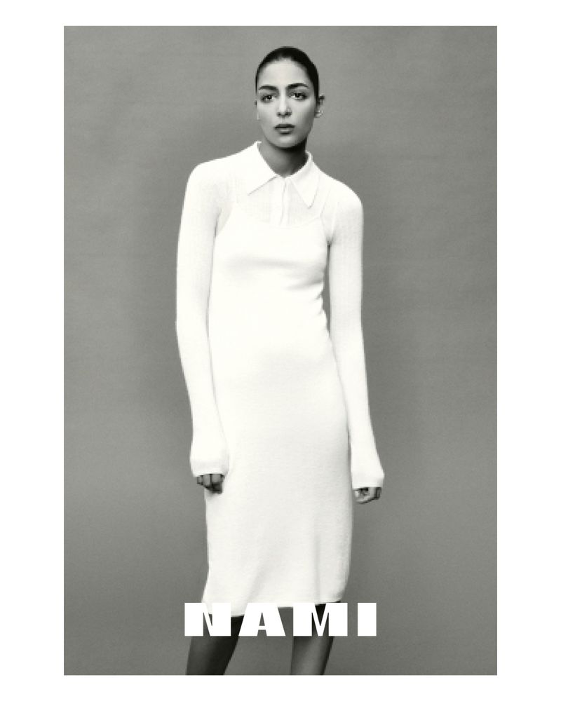 Monochrome shot of a woman posing indoors wearing a long-sleeve, collared dress. The text 'NAMI' is overlaid on the lower portion of the image.