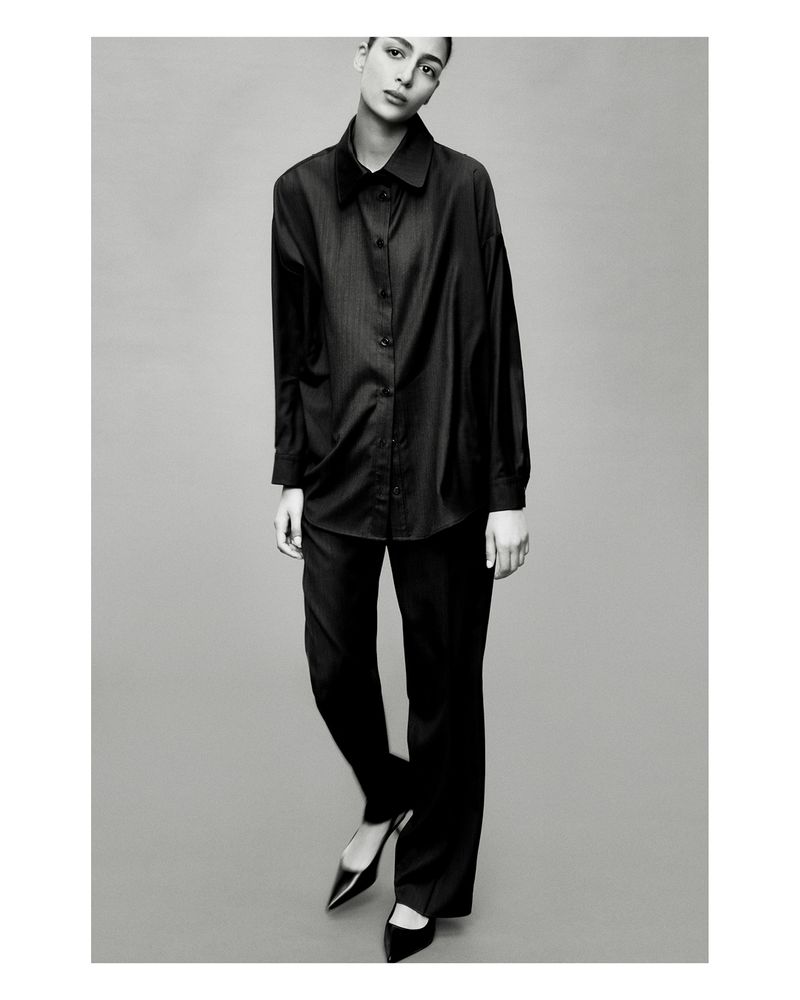 Black and white fashion shot of a woman in a black button-down shirt and black pants with heels, posing in front of a grey background.