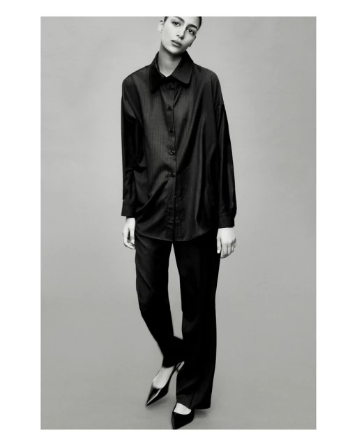 Black and white fashion shot of a woman in a black button-down shirt and black pants with heels, posing in front of a grey background.