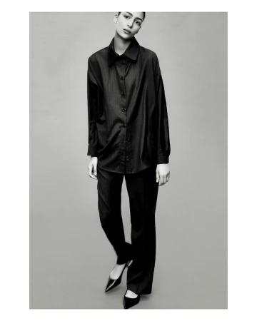 Black and white fashion shot of a woman in a black button-down shirt and black pants with heels, posing in front of a grey background.