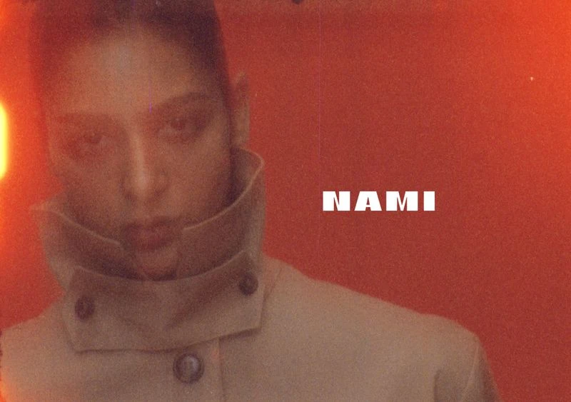 Close-up shot of an adult female model in a tan jacket posing against a reddish-orange background with the name 'NAMI' in white letters.