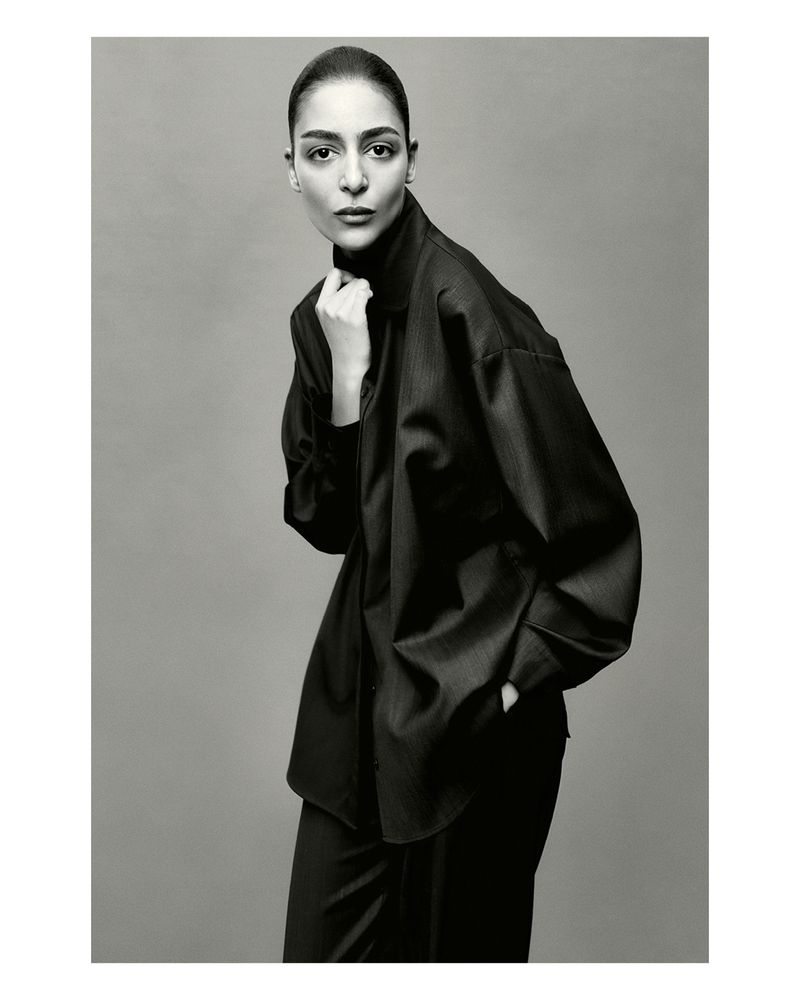 Monochrome portrait of a woman with dark hair wearing a dark, oversized collared shirt, posing in a photo studio with a neutral background.