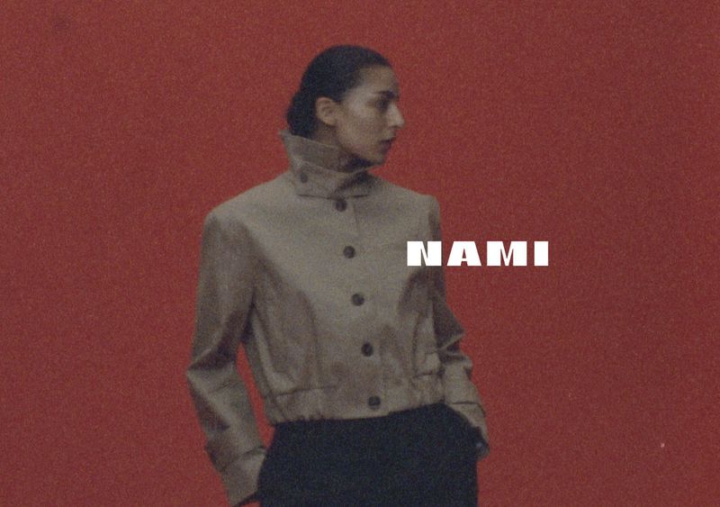 Woman in cropped jacket poses against red backdrop, featuring 'Nami' in bold text. Fashion editorial shot, studio lighting.