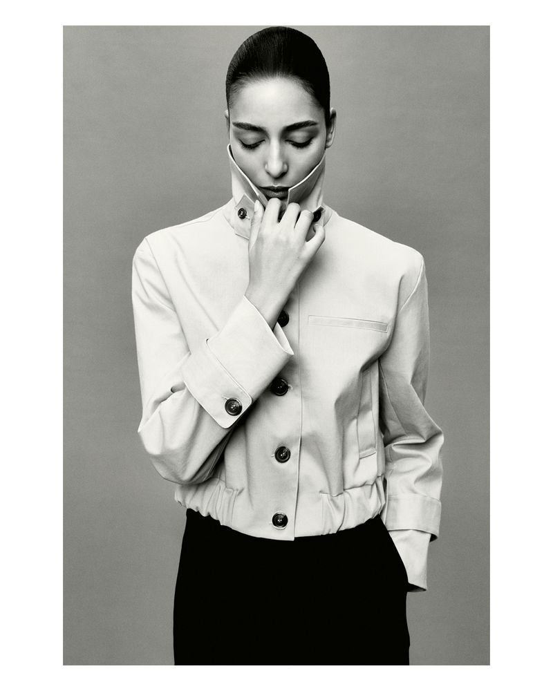 Monochrome portrait of a woman in a cream-colored jacket with her eyes closed and hand at her neck, against a light gray background.