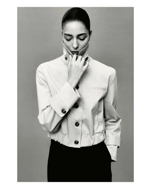 Monochrome portrait of a woman in a cream-colored jacket with her eyes closed and hand at her neck, against a light gray background.