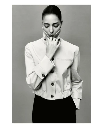 Monochrome portrait of a woman in a cream-colored jacket with her eyes closed and hand at her neck, against a light gray background.