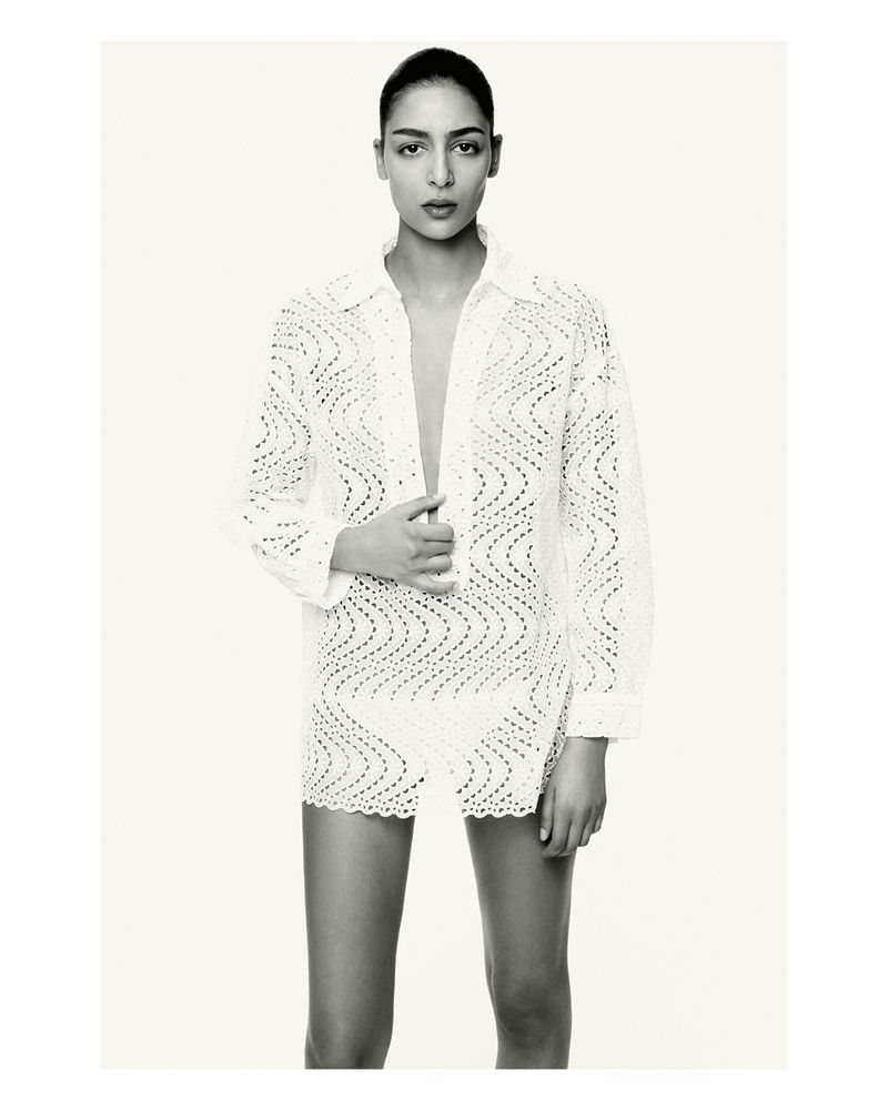 A woman models a white crochet mini dress with a matching button-down shirt, posing in a bright photo studio against a seamless backdrop.