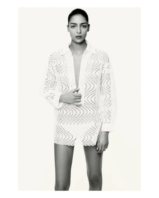 A woman models a white crochet mini dress with a matching button-down shirt, posing in a bright photo studio against a seamless backdrop.