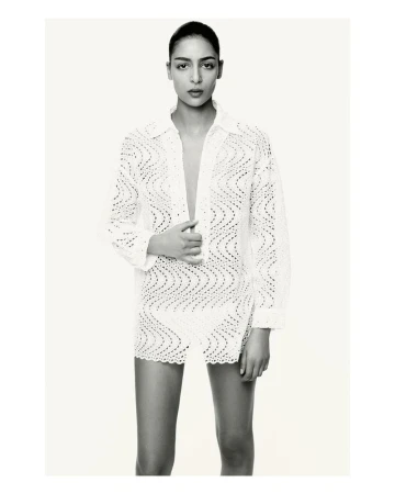 A woman models a white crochet mini dress with a matching button-down shirt, posing in a bright photo studio against a seamless backdrop.