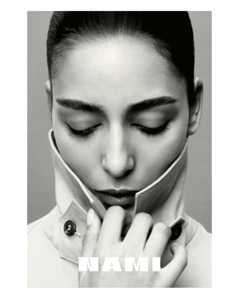 Monochrome headshot of an adult woman with her eyes closed, wearing a trench coat and posed in a bright, neutral-toned indoor setting.