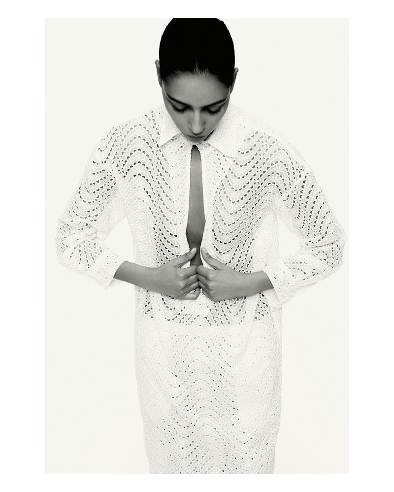 Black-and-white fashion shot of an adult woman with hands near the buttoned collar of a patterned, long-sleeved white shirt dress.