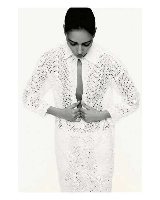 Black-and-white fashion shot of an adult woman with hands near the buttoned collar of a patterned, long-sleeved white shirt dress.