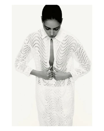 Black-and-white fashion shot of an adult woman with hands near the buttoned collar of a patterned, long-sleeved white shirt dress.