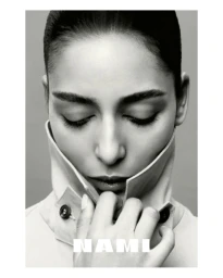 Close-up portrait of model Nora Attal wearing a collared coat with her hand held near her chin against a neutral grey background.