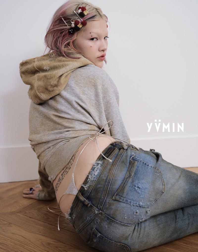 Young adult woman with rose-tinged hair, wearing a hoodie and distressed jeans, gazes back toward the camera indoors against a white wall.