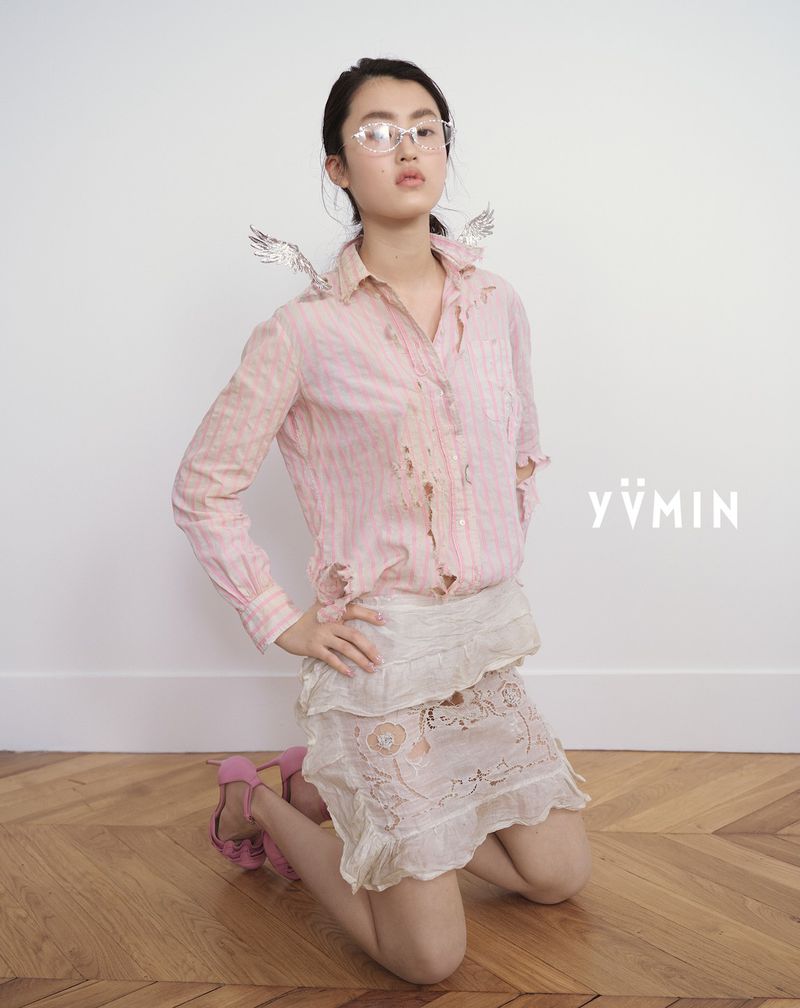 Young woman with silver wings kneeling in a studio wearing glasses, a pink striped shirt, a white skirt, and pink high heels.