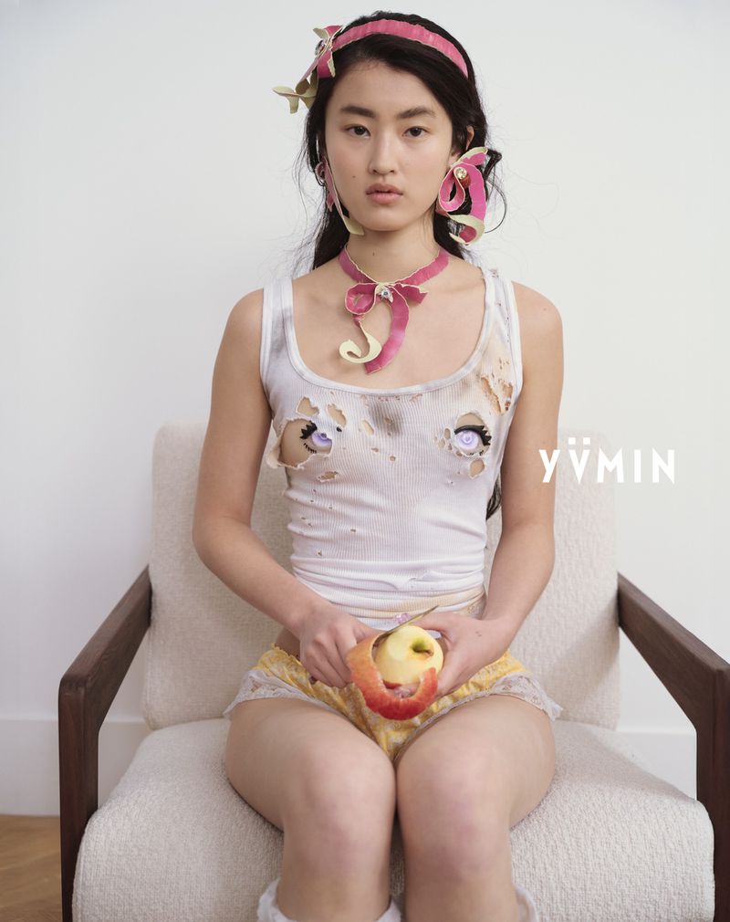 Woman with fair skin sits in a neutral chair, peeling an apple. She wears a white tank top with cartoon eyes and pink accessories.