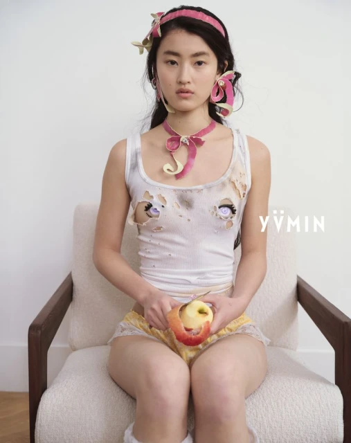 Woman with fair skin sits in a neutral chair, peeling an apple. She wears a white tank top with cartoon eyes and pink accessories.