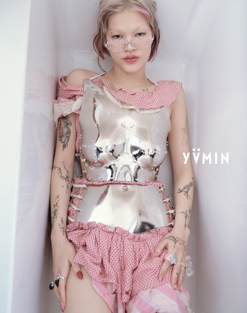 A female model with pink hair and transparent glasses wears a reflective breastplate and pink polka-dot skirt while posing indoors.