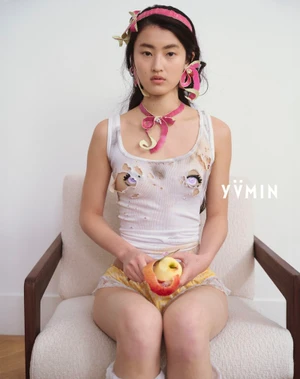Model seated on a beige armchair peeling an apple, wearing a distressed white tank top, pink ribbon choker, and yellow shorts.