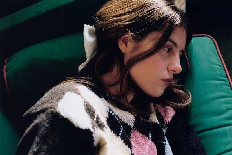 Young adult woman with brown hair reclines on green cushion wearing argyle sweater and white ribbon; portrait captures a blend of comfort and style.