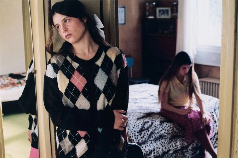 Two young women in a bedroom, one standing near a mirror wearing an argyle sweater, and one sitting on a bed with a patterned cover.