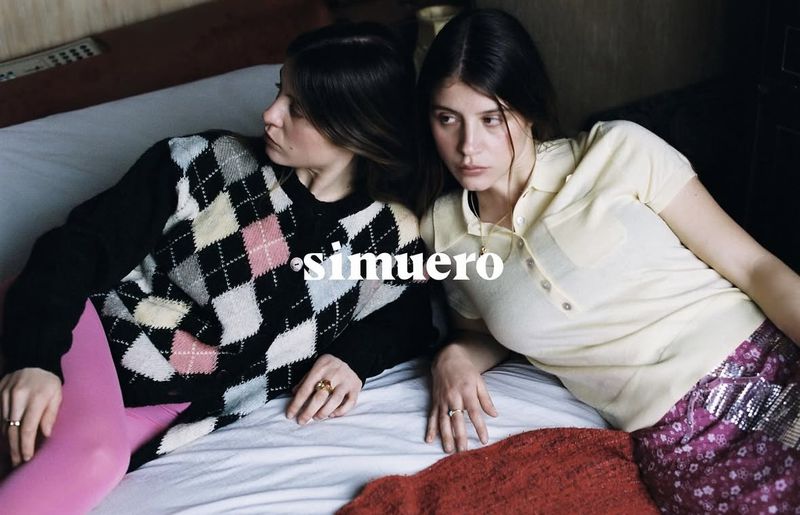 Two young women on a bed in a casual pose, one in a patterned sweater and pink tights, the other in a polo shirt and patterned skirt.