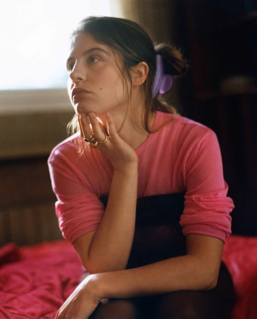 Portrait of a woman with brown hair pulled back with a hair clip, wearing a pink sweater, resting her chin on her hand indoors.
