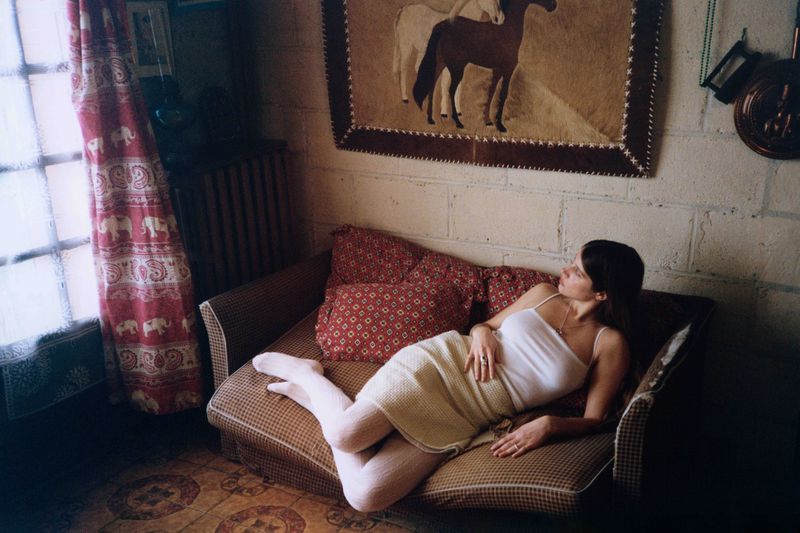 An adult woman relaxes on a couch in an indoor setting, bathed in soft natural light near a window, next to patterned pillows and curtains.
