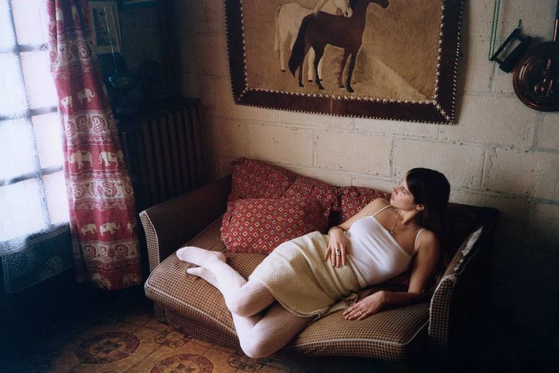 An adult woman relaxes on a couch in an indoor setting, bathed in soft natural light near a window, next to patterned pillows and curtains.