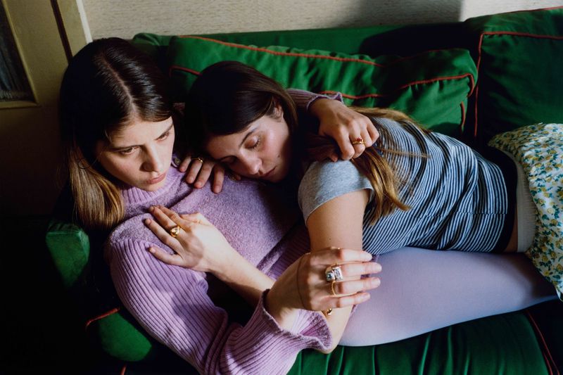 Two women embrace on a green couch in a relaxed indoor setting, highlighting their close relationship and casual attire, with a warm color palette.