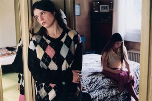 A young woman wearing an argyle cardigan in the foreground with another woman seated on a bed in the background of a domestic interior