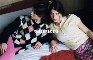 Two women lying on a bed, one wearing an argyle cardigan and pink trousers, the other wearing a yellow polo and patterned skirt