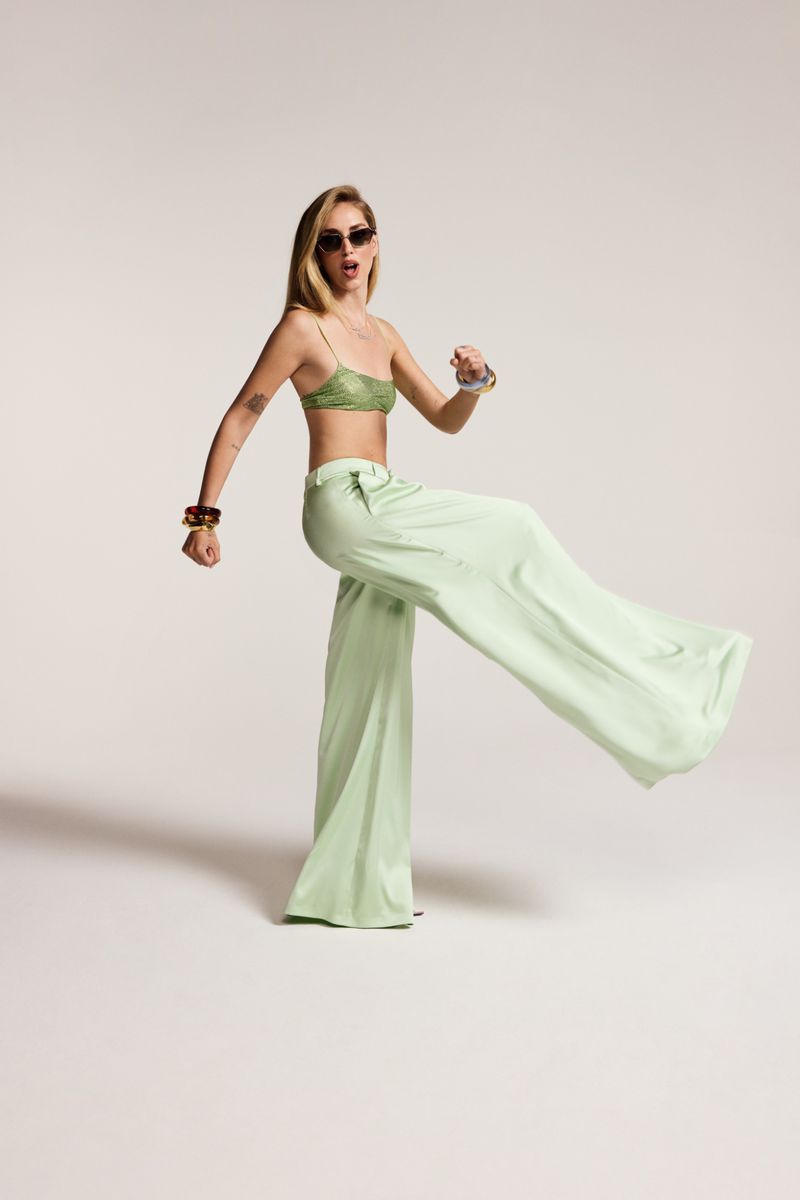 Adult female fashion model poses in pale green wide-leg pants and lace bra top against a neutral background with natural lighting.