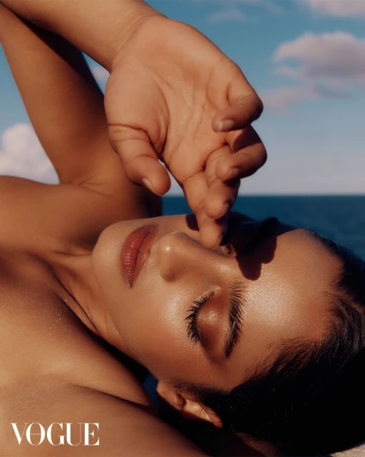 Close-up fashion shot of a tanned adult female model lying down with hand touching her nose, set against a blue sea and sky.