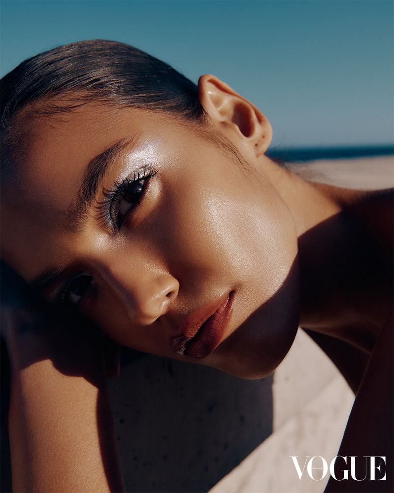 Close-up portrait of a young adult female fashion model with slicked back hair and dramatic eye makeup, posing in front of a beach.