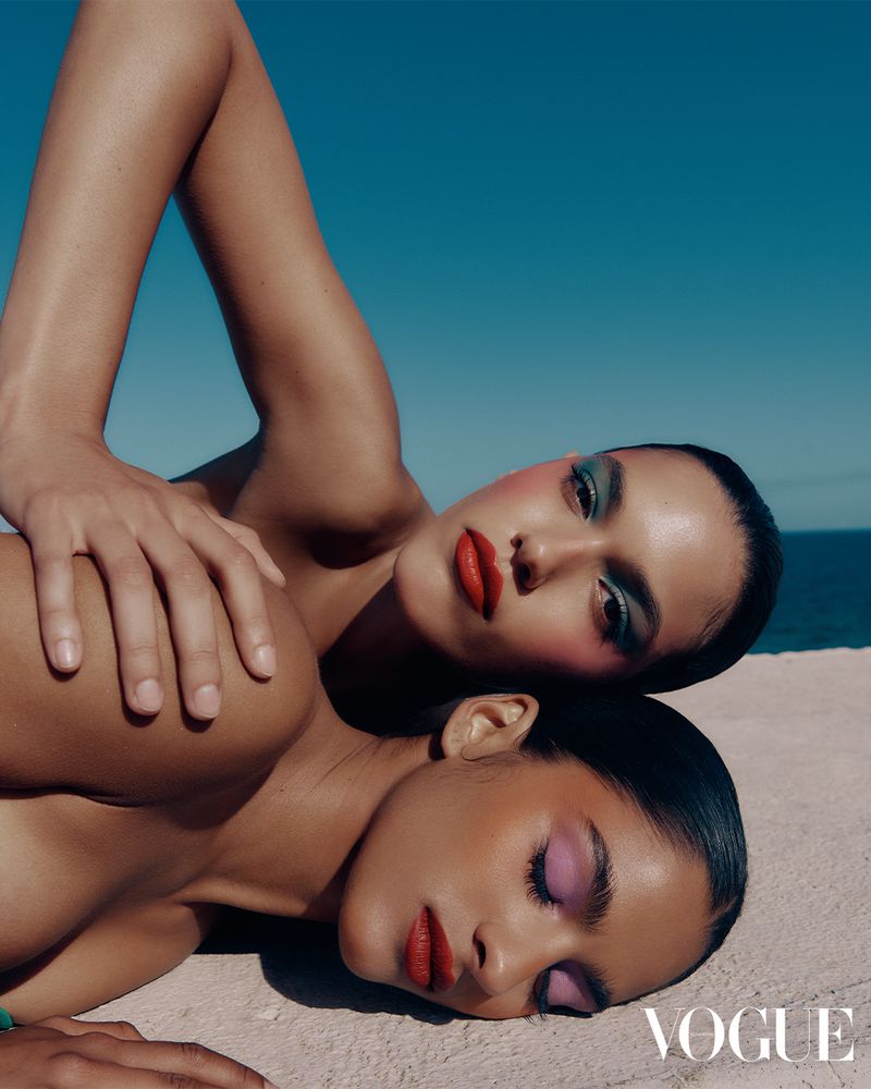 Two women pose on a beach against a clear blue sky. They wear bright makeup in a fashion editorial with a magazine logo visible.