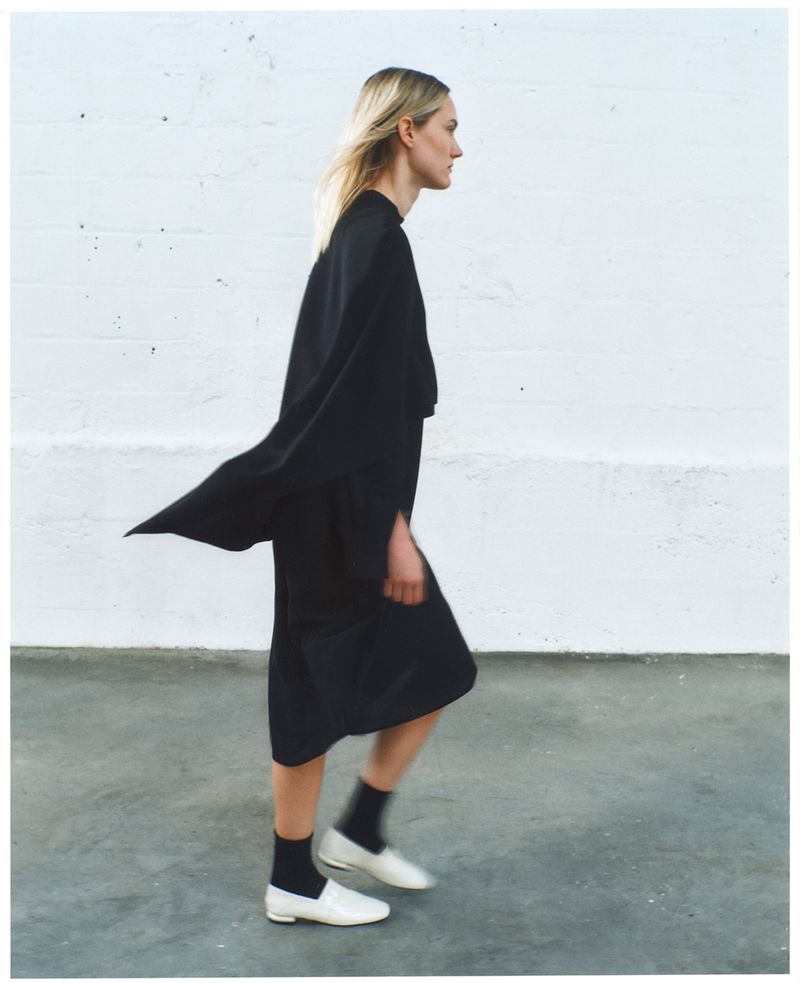 Woman walks in profile wearing a black outfit with white shoes against a white brick wall and grey floor, fashion shot, minimalist.