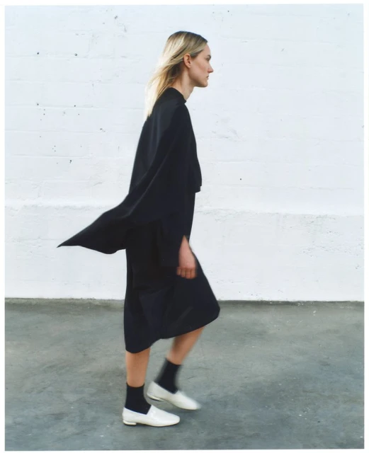 Woman walks in profile wearing a black outfit with white shoes against a white brick wall and grey floor, fashion shot, minimalist.