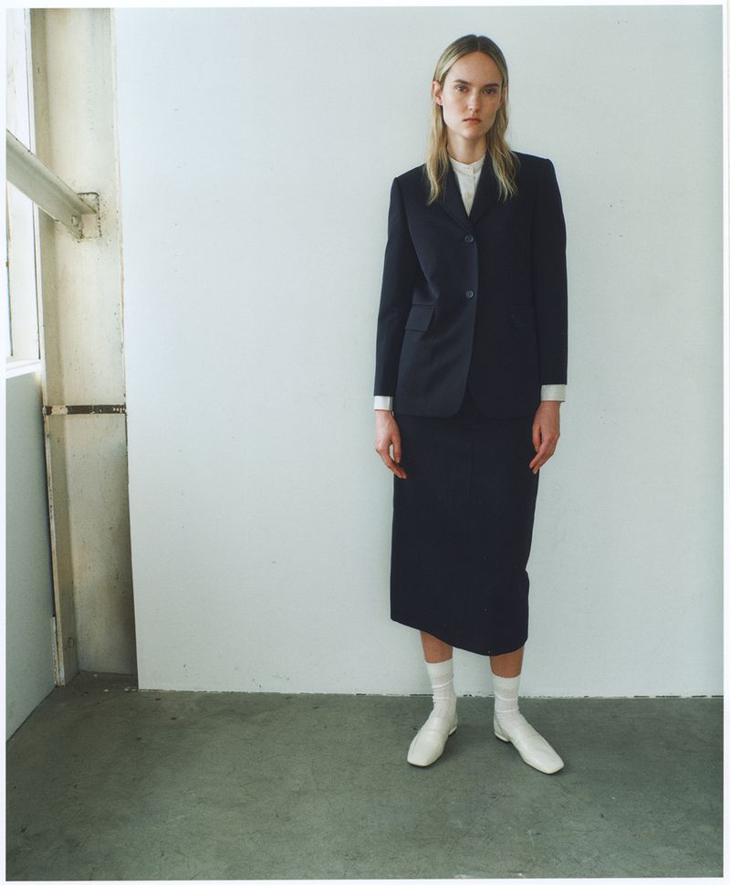A fair-skinned woman stands in a skirt suit, posed against a white wall near a window, with natural light enhancing the minimalist fashion aesthetic.