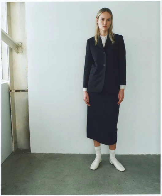 A fair-skinned woman stands in a skirt suit, posed against a white wall near a window, with natural light enhancing the minimalist fashion aesthetic.