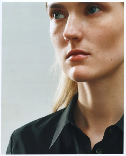 Close-up portrait of a blonde woman with blue eyes wearing a black collared shirt against a light grey background in natural lighting.