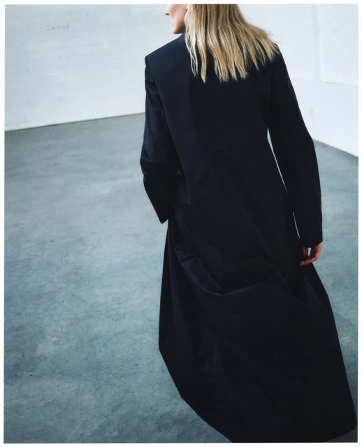 Back view of a woman with blonde hair wearing a long black coat indoors against a gray background, showcasing fashion and style.