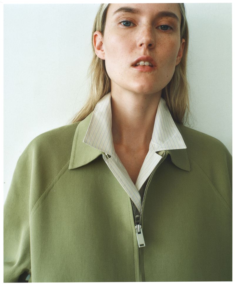 Close-up shot of a young woman with blonde hair wearing an olive green jacket and striped collared shirt against a light background.