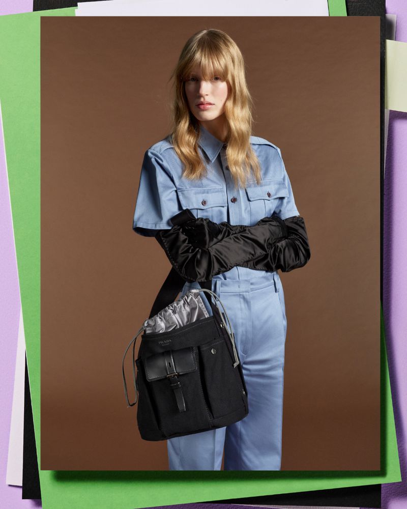 Fashion shot of a woman in light blue matching shirt and pants with a black Prada purse, black gloves, and a neutral backdrop.
