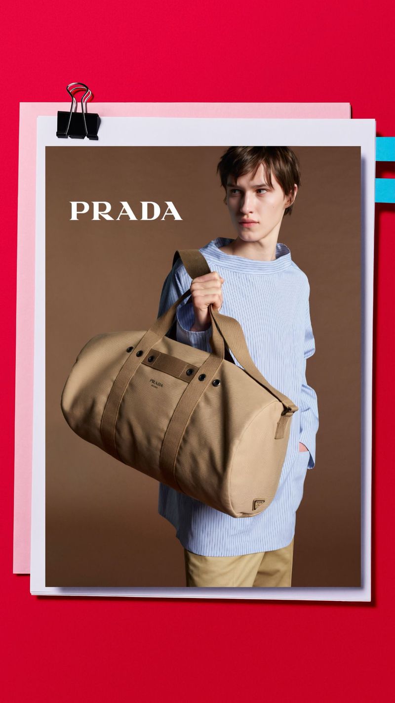 A person holding a khaki Prada duffel bag against a brown backdrop, framed by a red and pink border with a silver paper clip.
